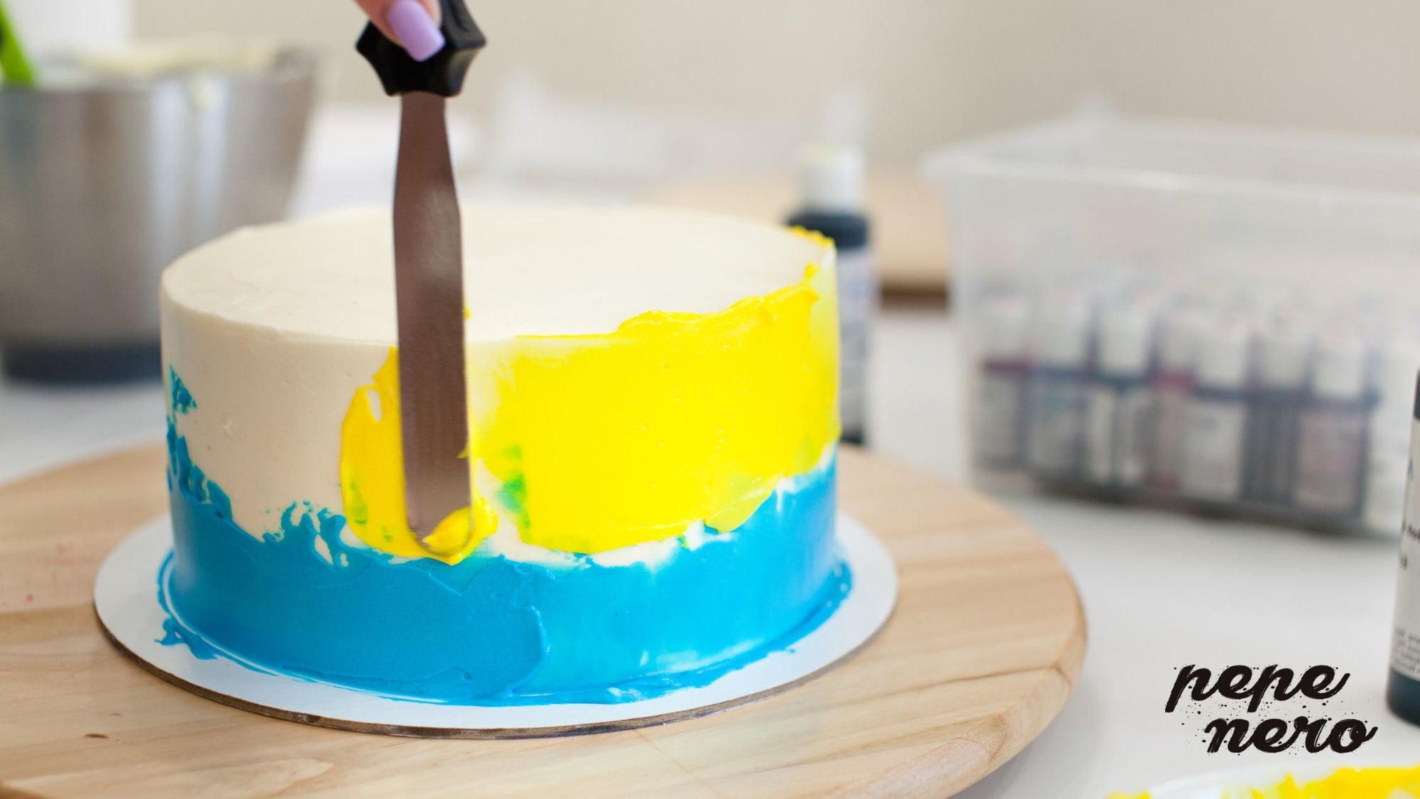 A cake with white icing being decorated with blue and yellow icing.