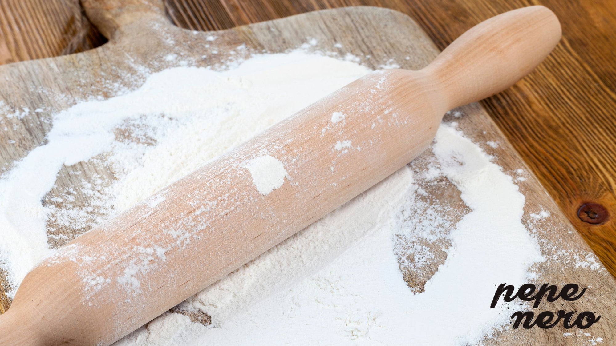 A wooden rolling pin that is lying in flour on a wooden chopping board